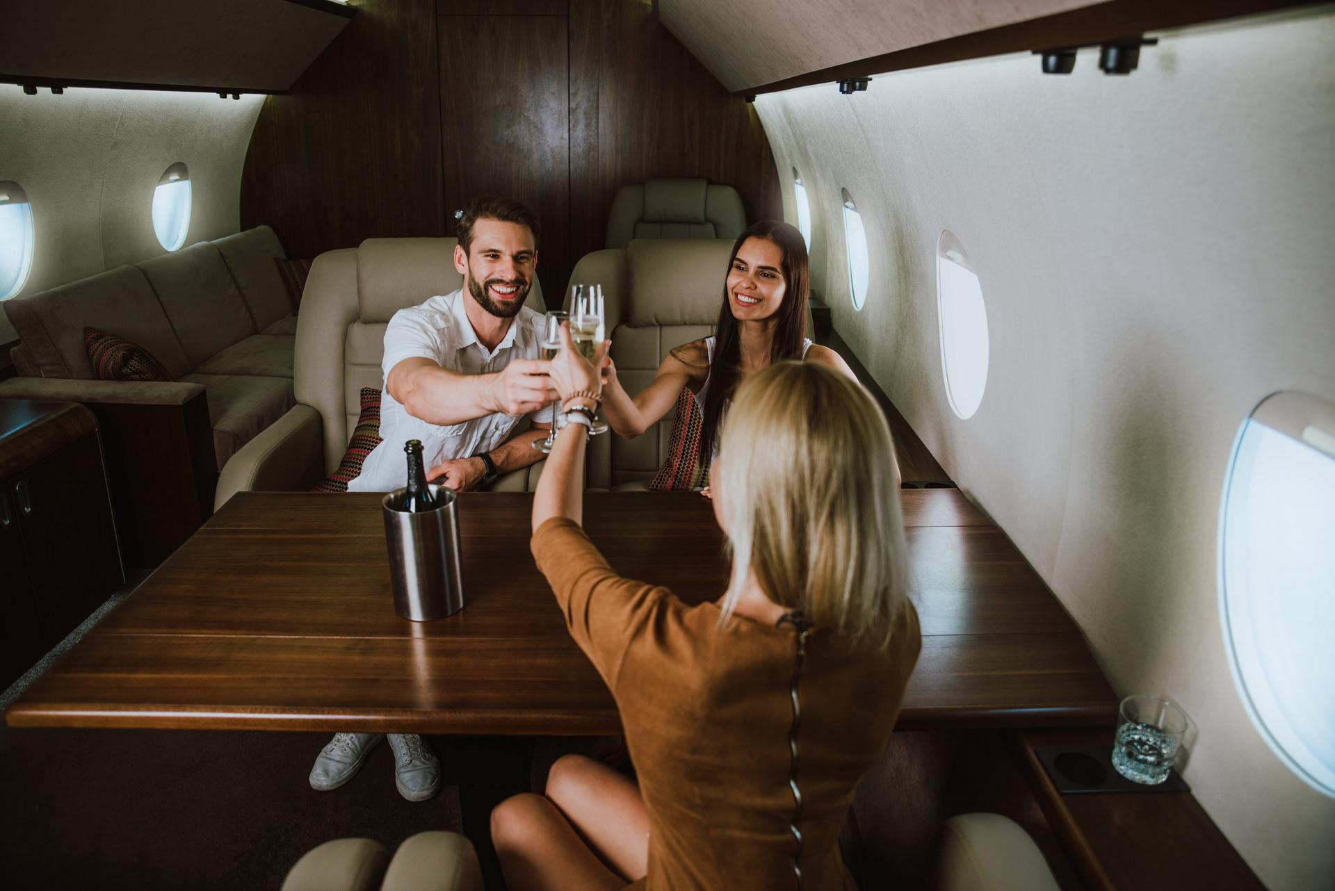 Three people sitting in a private jet, toasting with champagne glasses. A bottle of champagne in an ice bucket is on the table, courtesy of Premium Concierge Services.